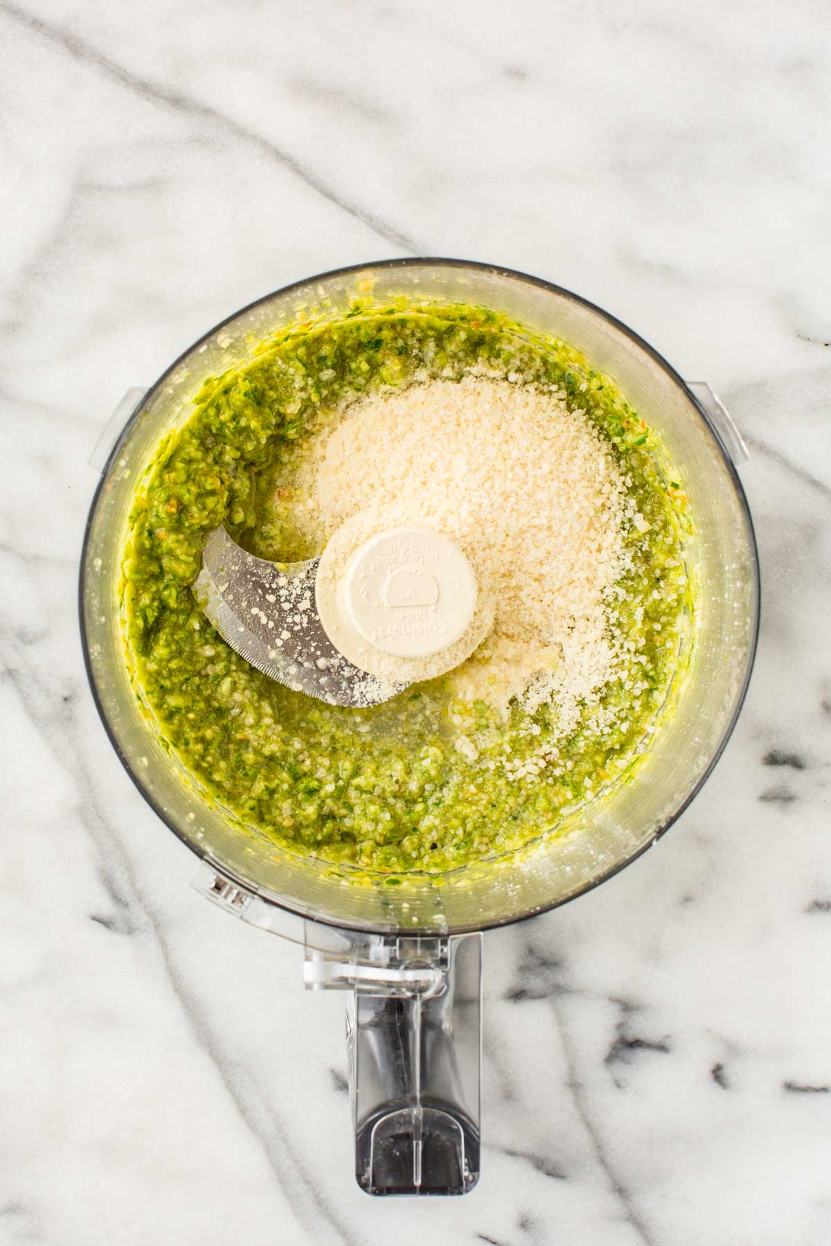 Bright green pesto mixture topped with finely grated Parmesan cheese in a food processor bowl mid-recipe.