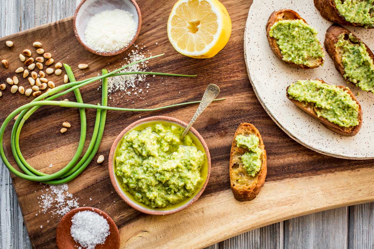 Rustic wooden board with crostini topped with garlic scape pesto, surrounded by pine nuts, grated cheese, lemon, and fresh garlic scapes.