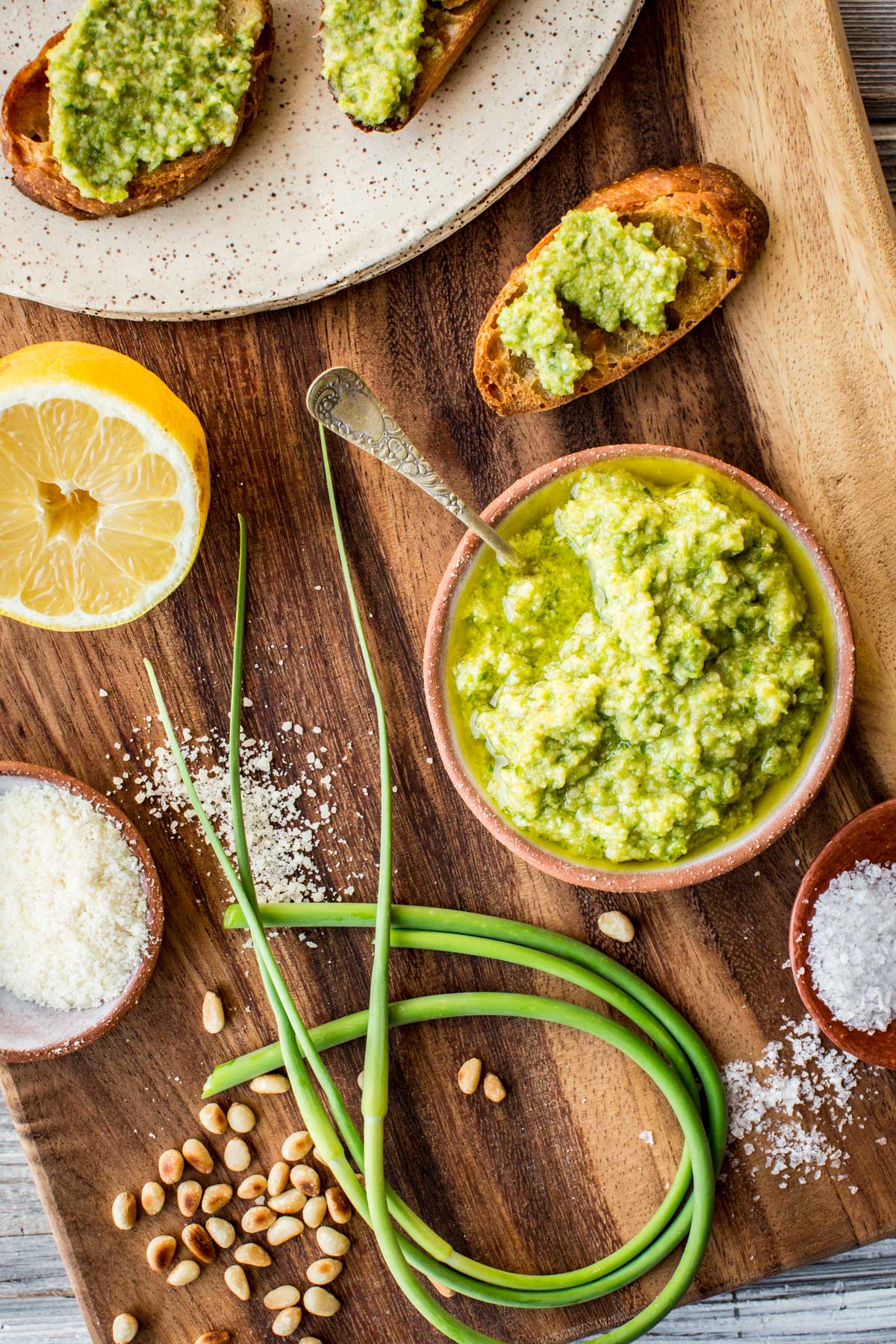 Close-up top view of pesto crostini on a speckled plate next to a bowl of pesto, lemon, sea salt, and raw garlic scapes.