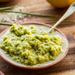 Close-up shot of creamy, textured garlic scape pesto in a ceramic bowl with a vintage spoon, surrounded by fresh ingredients.