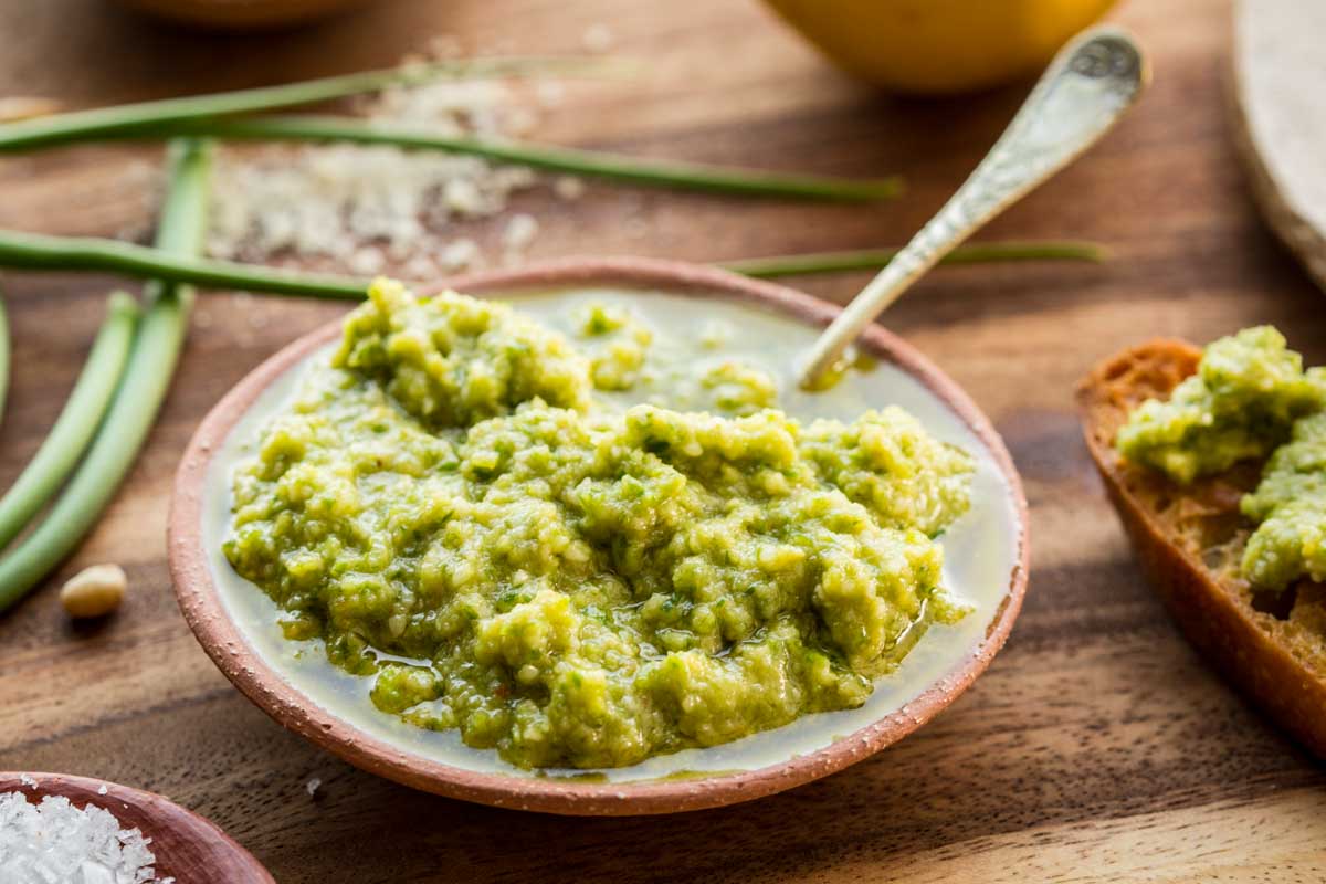 Close-up shot of creamy, textured garlic scape pesto in a ceramic bowl with a vintage spoon, surrounded by fresh ingredients.