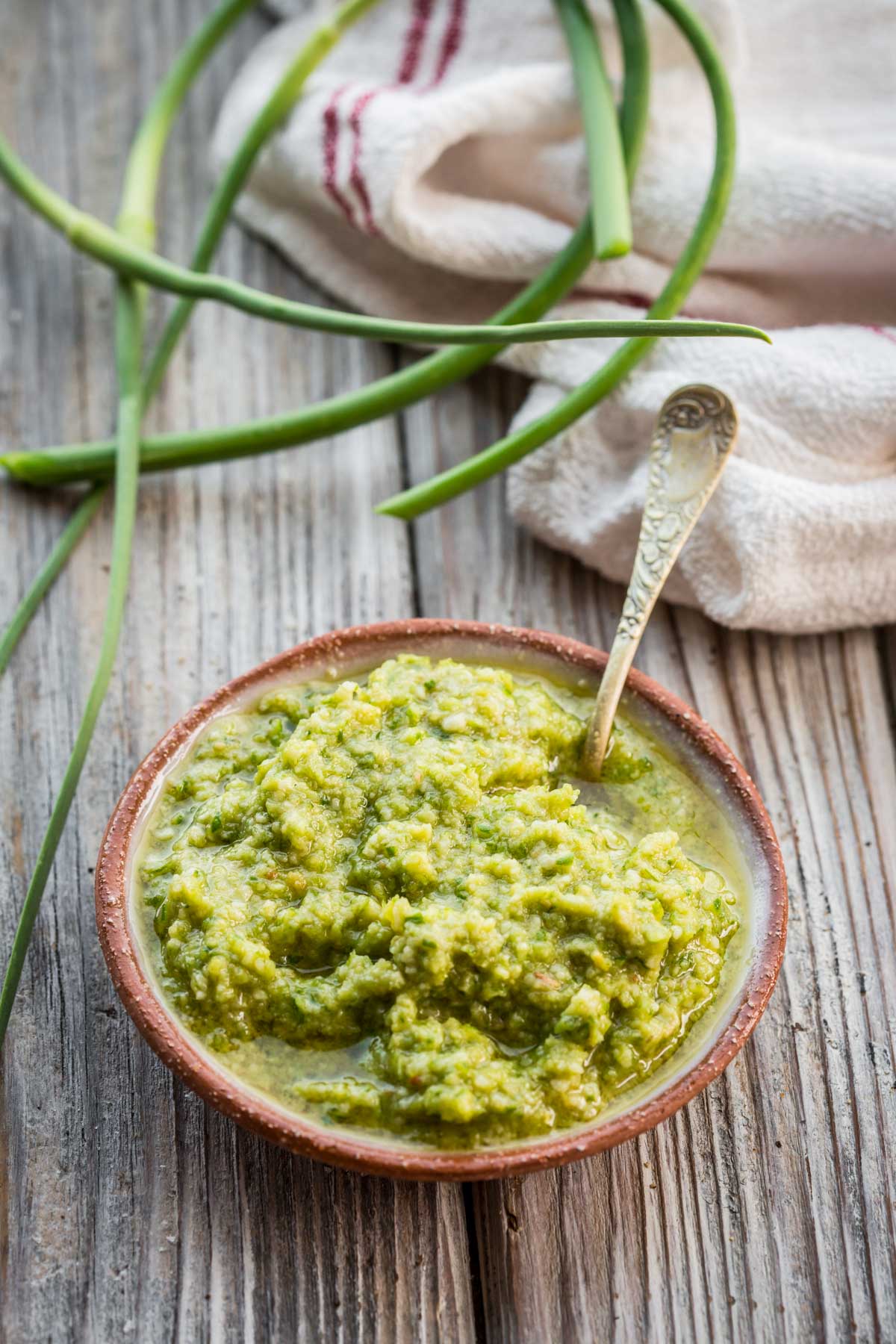 Vertical image of freshly made garlic scape pesto in a terracotta bowl with long green garlic scapes curling beside it.