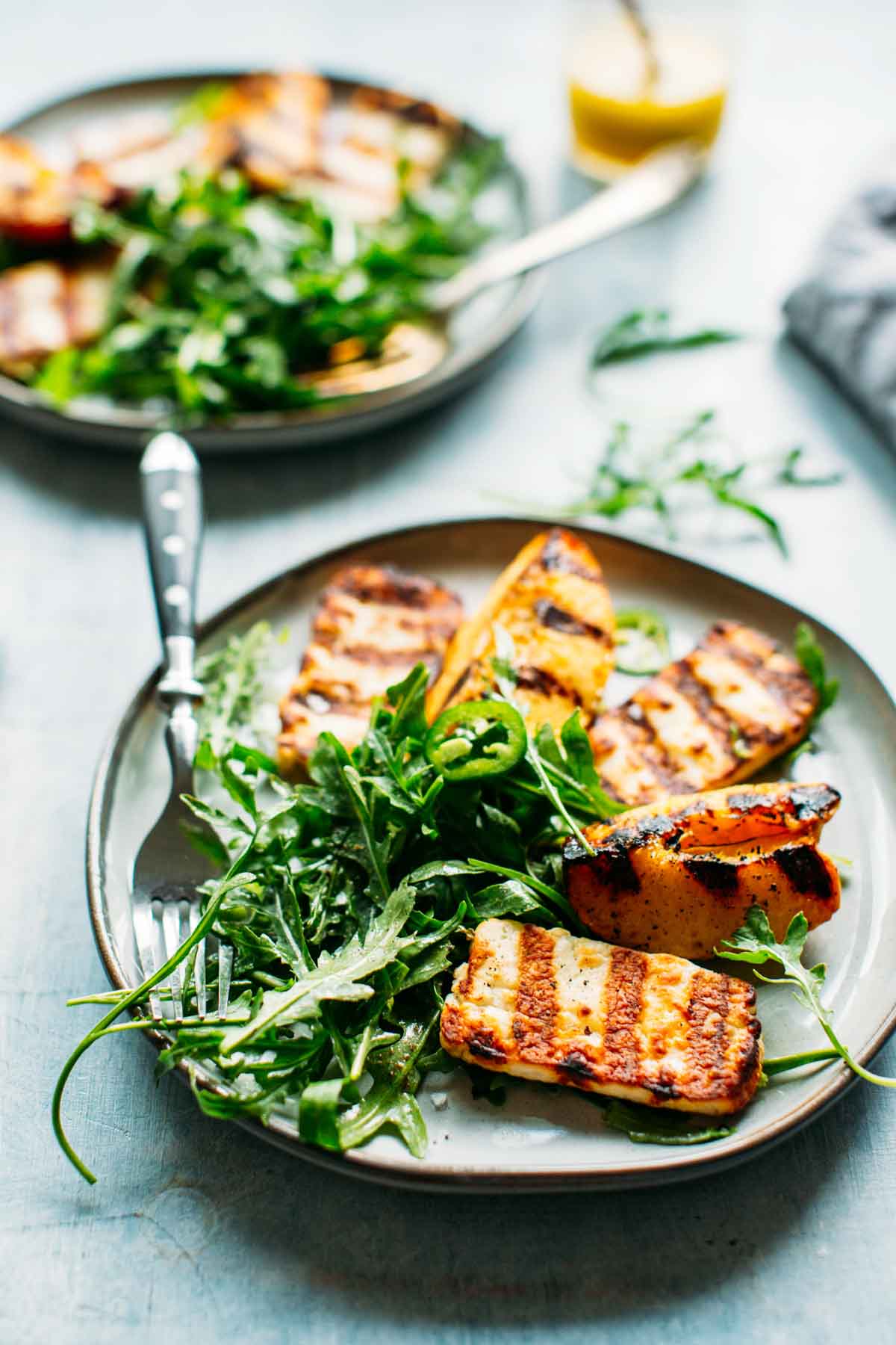 Plate of grilled halloumi and peaches over arugula, garnished with jalapeño slices, with a fork and vinaigrette jar in the background.