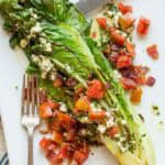 A grilled romaine lettuce half is plated with a generous sprinkling of crumbled blue cheese, chopped tomatoes, and crispy bacon bits. The charred edges of the romaine give it a smoky appearance, and the salad is drizzled with creamy blue cheese dressing. A fork and knife rest on the white plate.