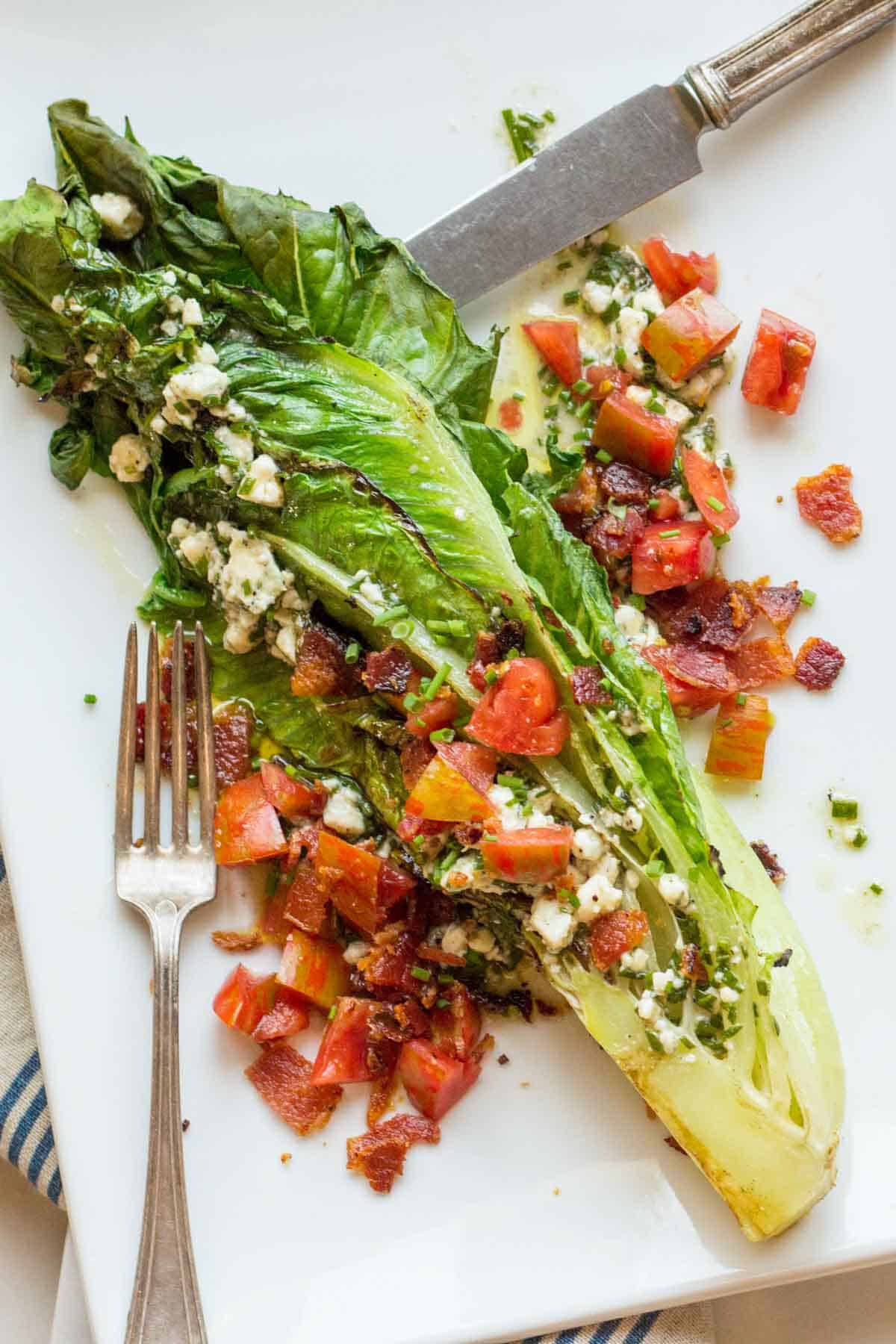 A grilled romaine lettuce half is plated with a generous sprinkling of crumbled blue cheese, chopped tomatoes, and crispy bacon bits. The charred edges of the romaine give it a smoky appearance, and the salad is drizzled with creamy blue cheese dressing. A fork and knife rest on the white plate.