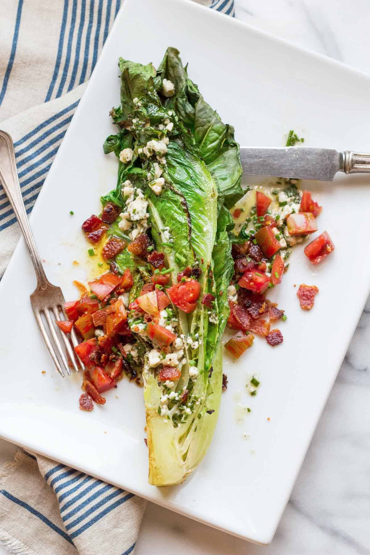 A close-up top-down view of a grilled romaine wedge salad topped with diced tomatoes, crispy bacon pieces, and blue cheese crumbles, dressed with a creamy vinaigrette. The lettuce is slightly charred and plated on a white rectangular dish with vintage silverware and a striped cloth napkin.