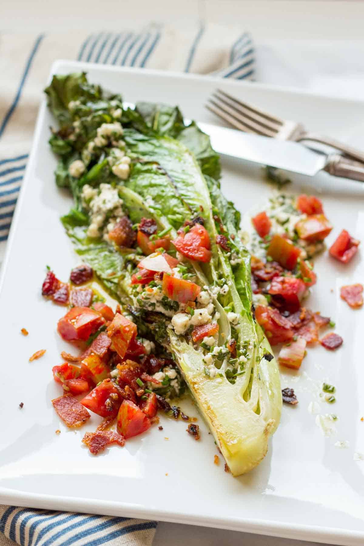 Grilled romaine lettuce is served whole with char marks visible on the leaves. It's topped with a chunky mixture of tomatoes, bacon, and blue cheese dressing. The salad is arranged diagonally across a white plate with a knife placed to the right and a fork to the left.