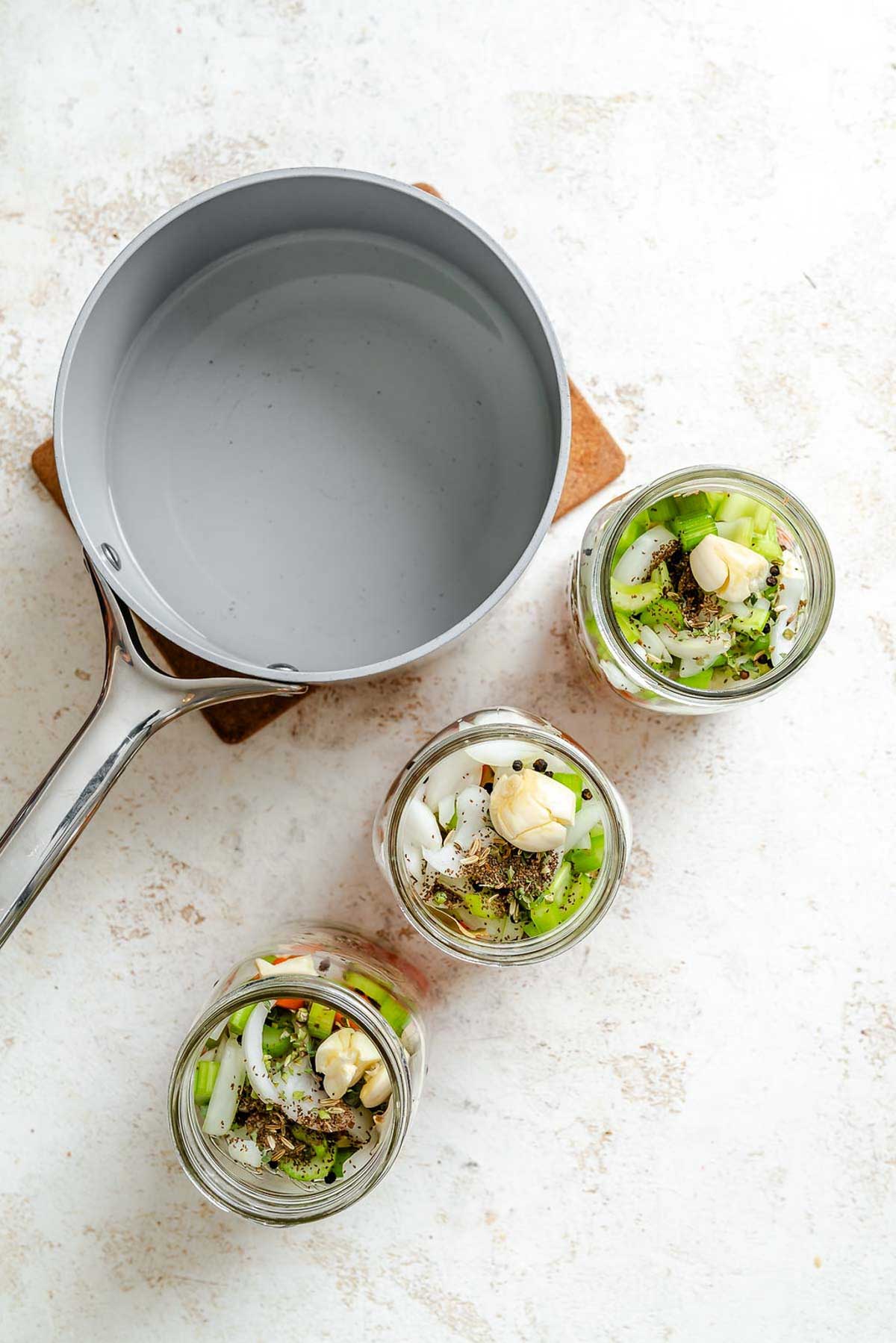Jars filled with seasoned vegetables ready for brine – overhead shot of garlic, herbs, and chopped veggies in glass jars next to pot of liquid.