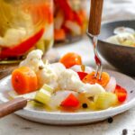 Close-up of a fork lifting a slice of pickled carrot from a plate of colorful giardiniera – includes cauliflower, red pepper, celery, and onion, lightly dressed in brine with visible herbs and peppercorns.