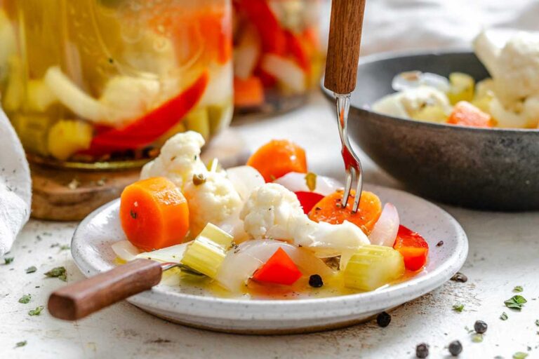 Close-up of a fork lifting a slice of pickled carrot from a plate of colorful giardiniera – includes cauliflower, red pepper, celery, and onion, lightly dressed in brine with visible herbs and peppercorns.