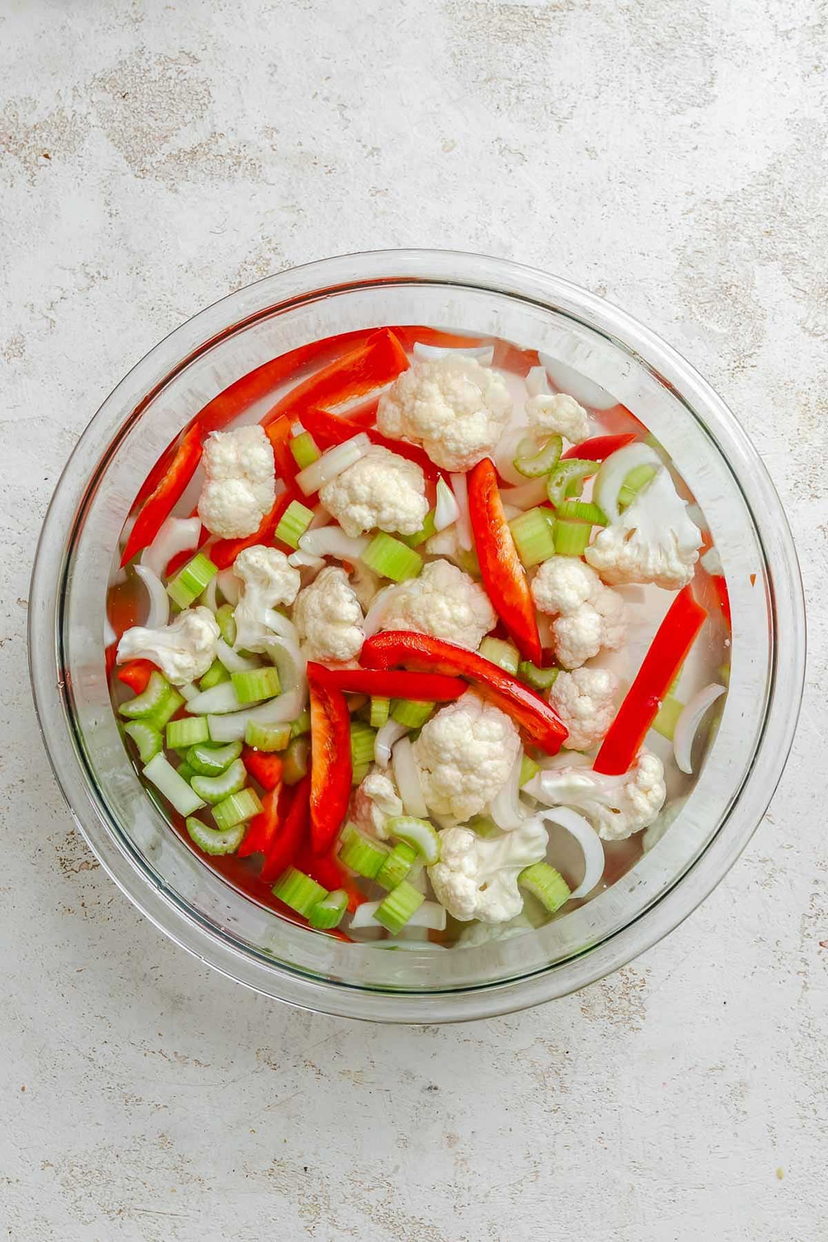 Overhead view of chopped raw vegetables in a glass bowl soaking in a brine – shows cauliflower florets, celery slices, red bell pepper strips, and onion.