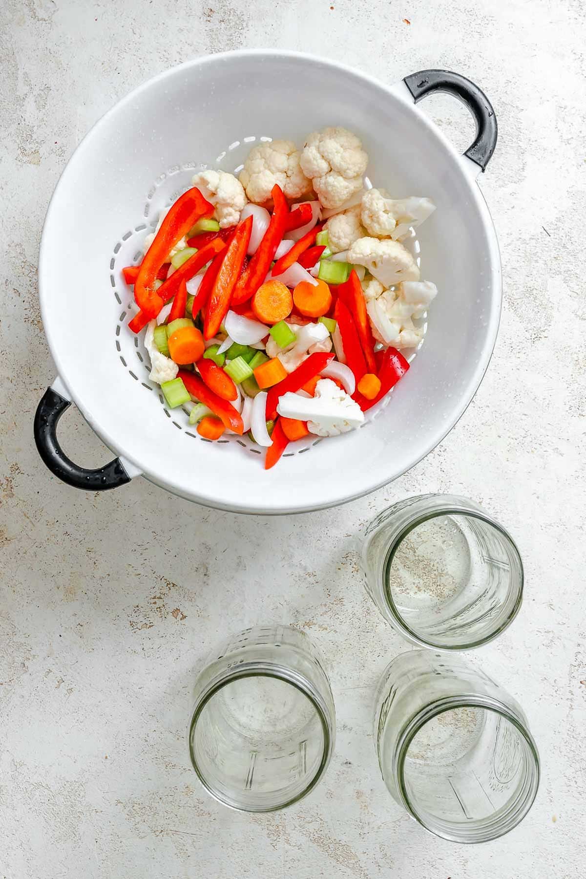 Strained giardiniera vegetables in a white colander with black handles – vibrant mix of chopped red peppers, carrots, celery, onions, and cauliflower.