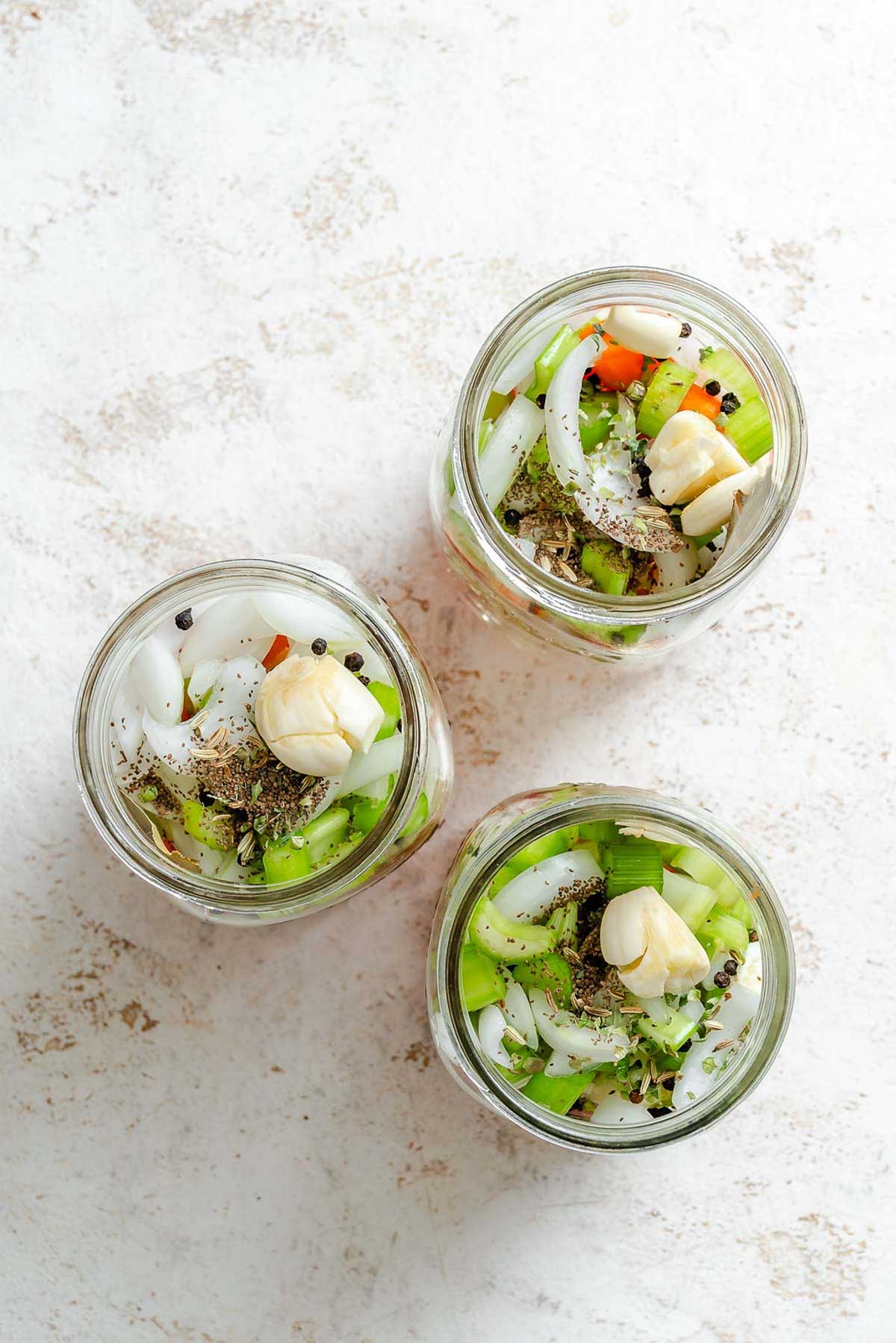 Close-up of filled jars topped with garlic cloves and seasoning – fresh giardiniera vegetables layered with whole garlic cloves, black peppercorns, and dried herbs.