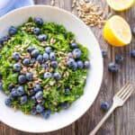A close-up of a white bowl filled with chopped kale, fresh blueberries, and sunflower seeds, set on a rustic wooden table. A halved lemon and scattered seeds and berries add visual interest around the bowl.