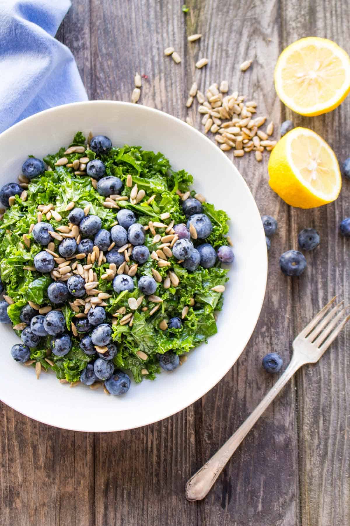 A close-up of a white bowl filled with chopped kale, fresh blueberries, and sunflower seeds, set on a rustic wooden table. A halved lemon and scattered seeds and berries add visual interest around the bowl.