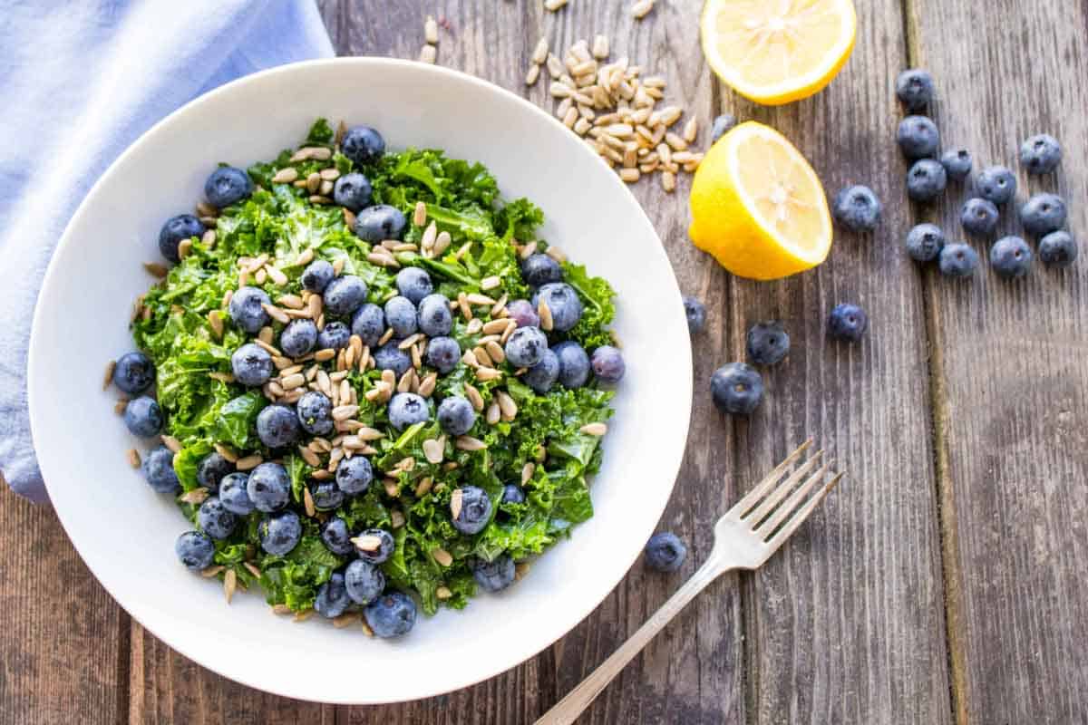 A wide shot of the kale and blueberry salad in a white bowl, topped with sunflower seeds, surrounded by lemon halves, loose blueberries, and sunflower seeds scattered on a wooden table, creating a fresh, natural scene.