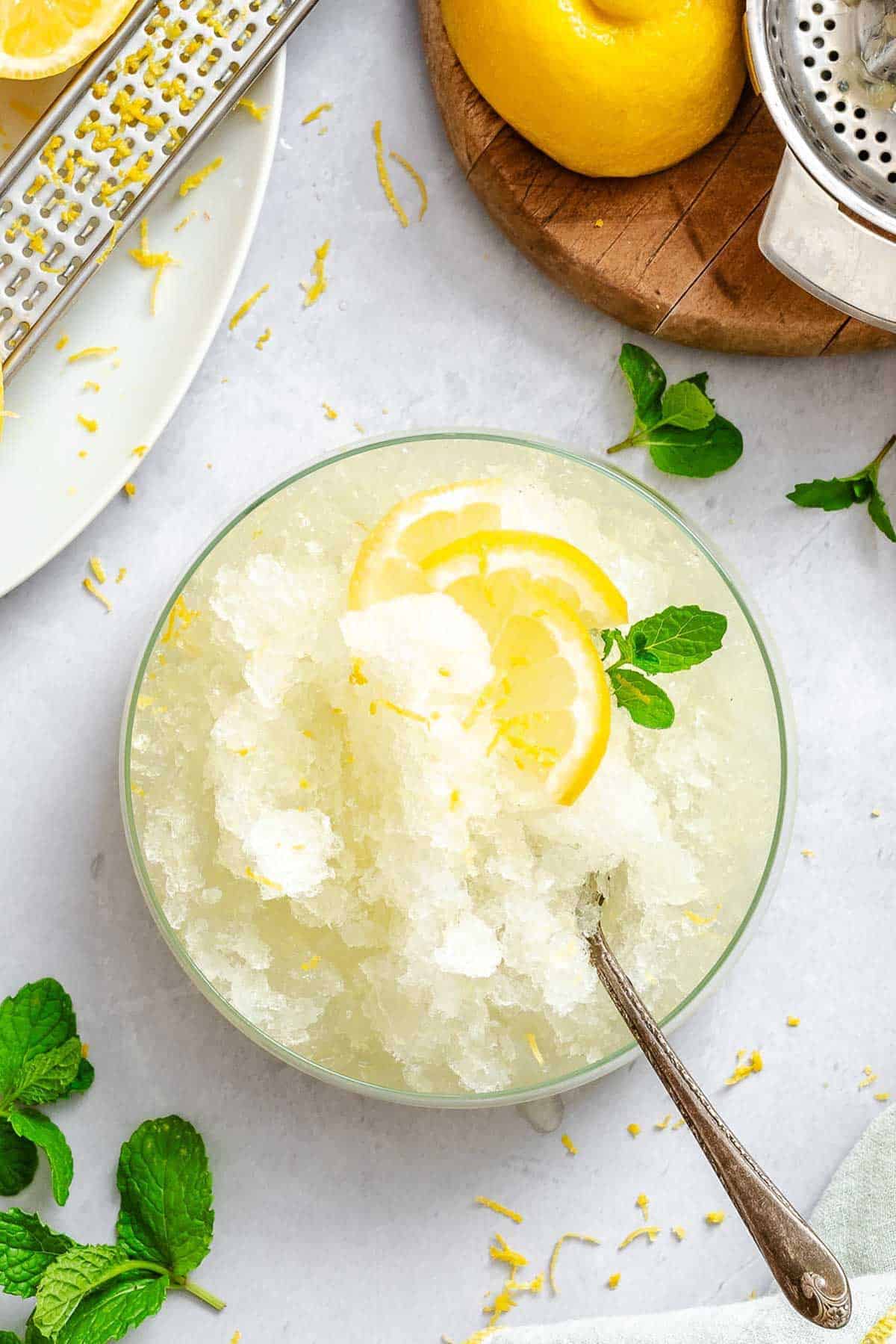 A glass bowl filled with icy lemon granita topped with thin lemon slices and a sprig of mint, with lemon zest and mint leaves scattered around on a light surface.
