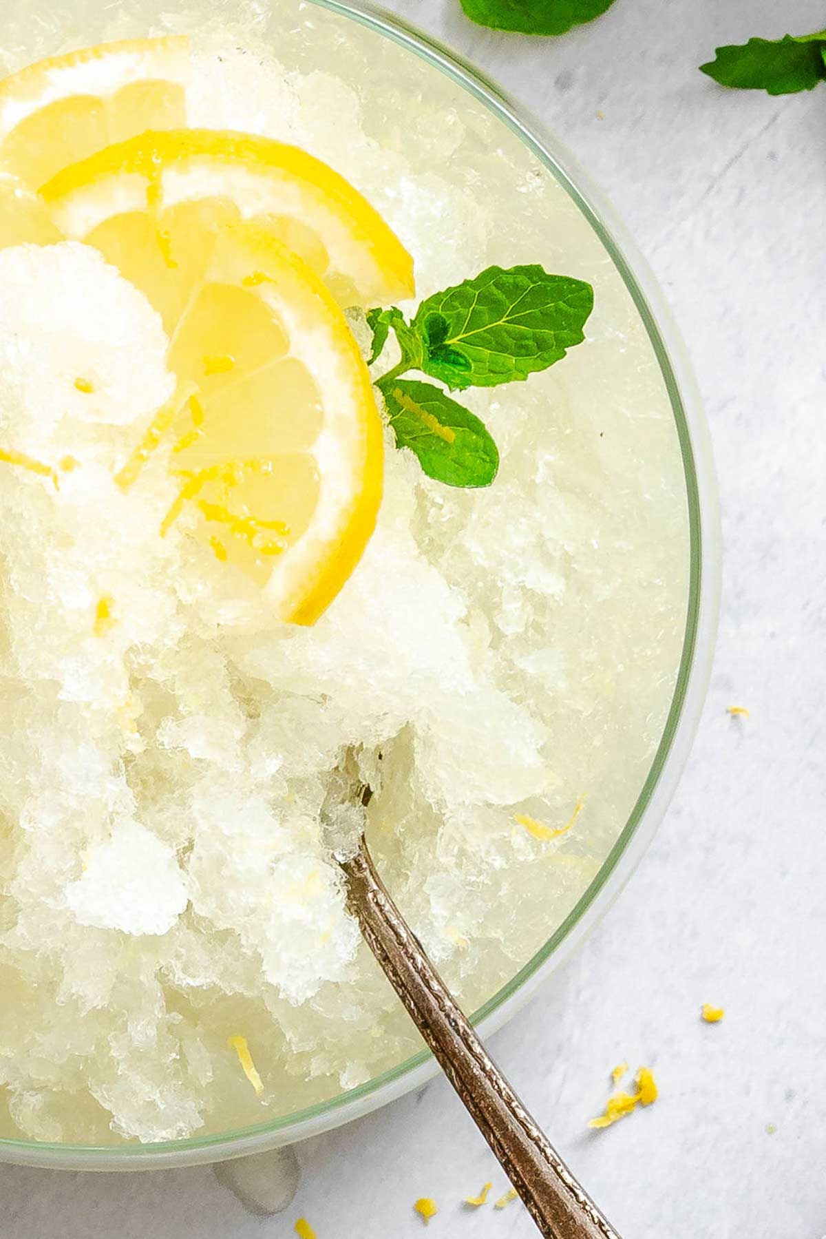 Close-up of lemon granita in a glass bowl with a spoon, garnished with lemon slices and a sprig of mint; crushed ice glistens against the pale yellow background.