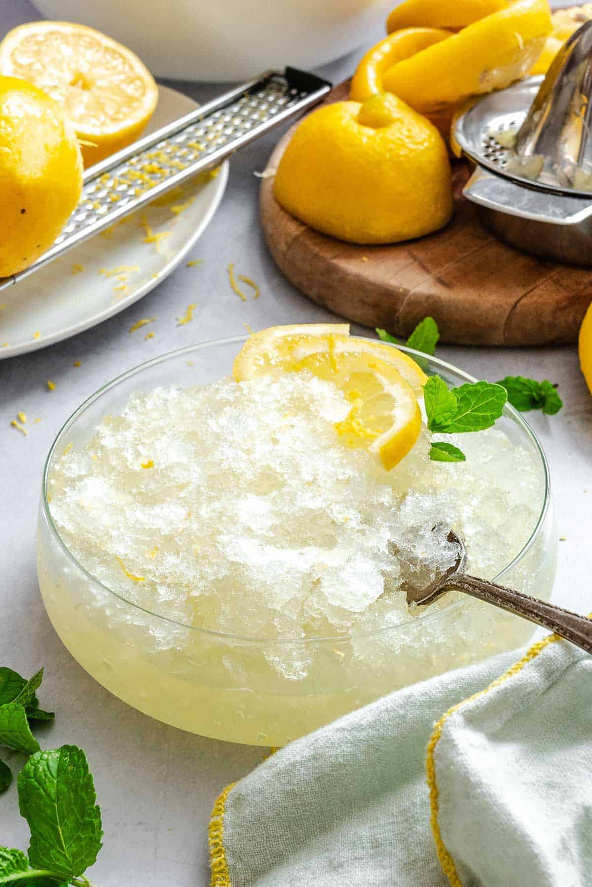 Angled view of a bowl of lemon granita placed on a table with a kitchen towel, surrounded by squeezed lemon halves, mint leaves, and a citrus juicer in the background.