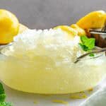 Side view of a frosty lemon granita mound in a glass bowl, garnished with lemon wedges and mint, with whole and halved lemons in the background.