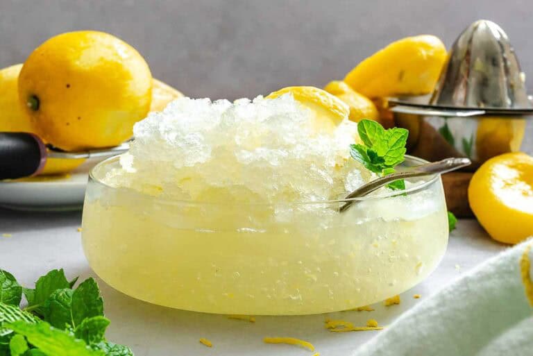 Side view of a frosty lemon granita mound in a glass bowl, garnished with lemon wedges and mint, with whole and halved lemons in the background.