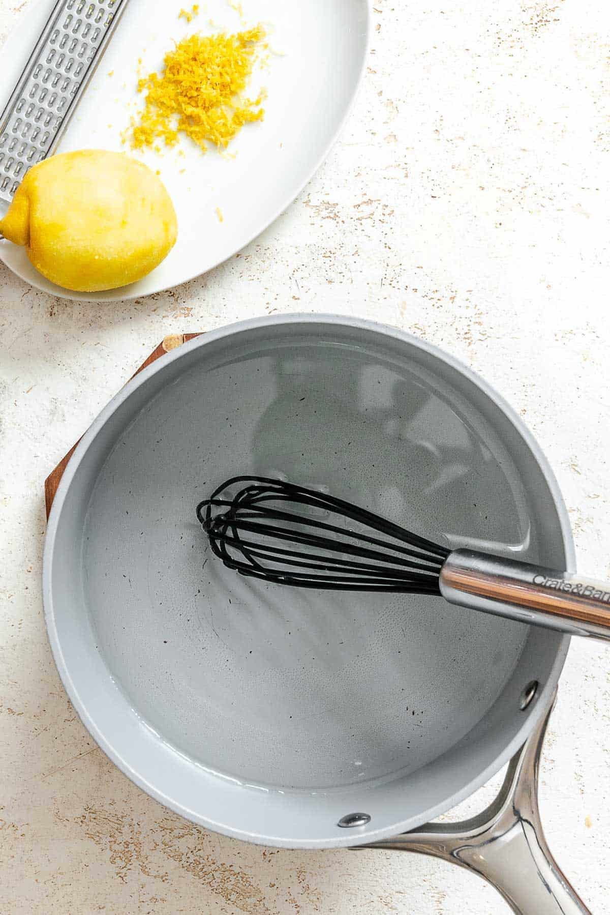A saucepan of clear water with a whisk, placed on a textured countertop with a zested lemon and pile of lemon zest nearby, showing preparation beginnings.
