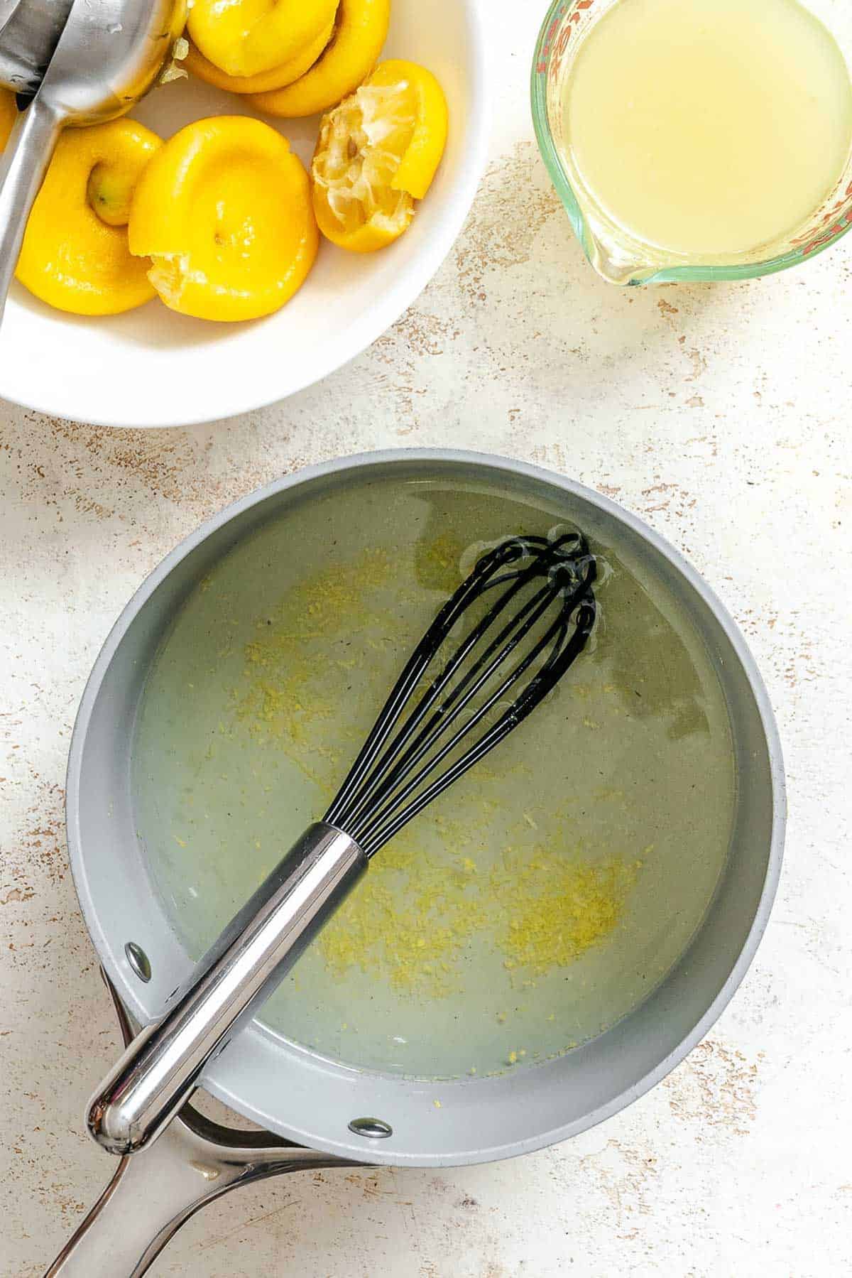 A saucepan filled with lemon zest-infused syrup, whisk inside, with a bowl of squeezed lemon halves and a measuring cup of juice nearby.