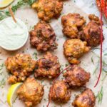 Overhead view of golden-brown lobster corn fritters scattered on newspaper-lined parchment with fresh tarragon sprigs, lemon wedges, and a small bowl of green tarragon aioli.