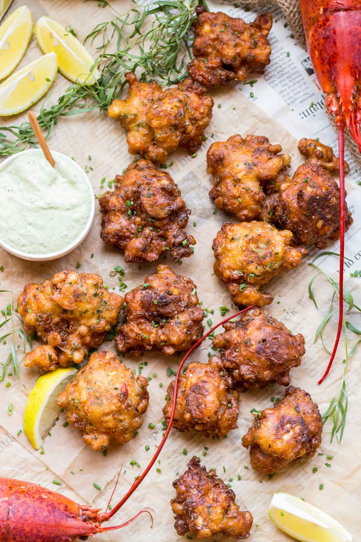 Overhead view of golden-brown lobster corn fritters scattered on newspaper-lined parchment with fresh tarragon sprigs, lemon wedges, and a small bowl of green tarragon aioli.