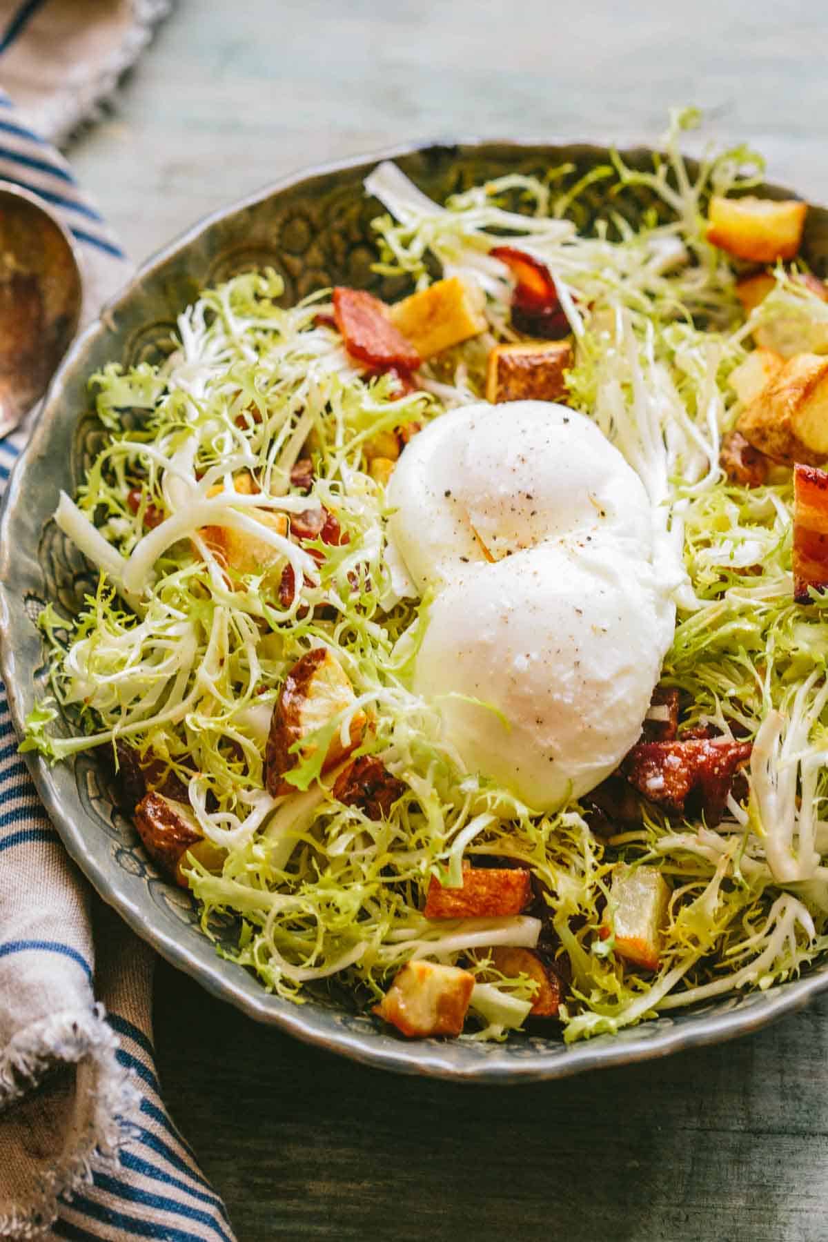 A bowl filled with frisée lettuce, crispy bacon lardons, golden roasted potatoes, and two perfectly poached eggs, seasoned with freshly ground black pepper.