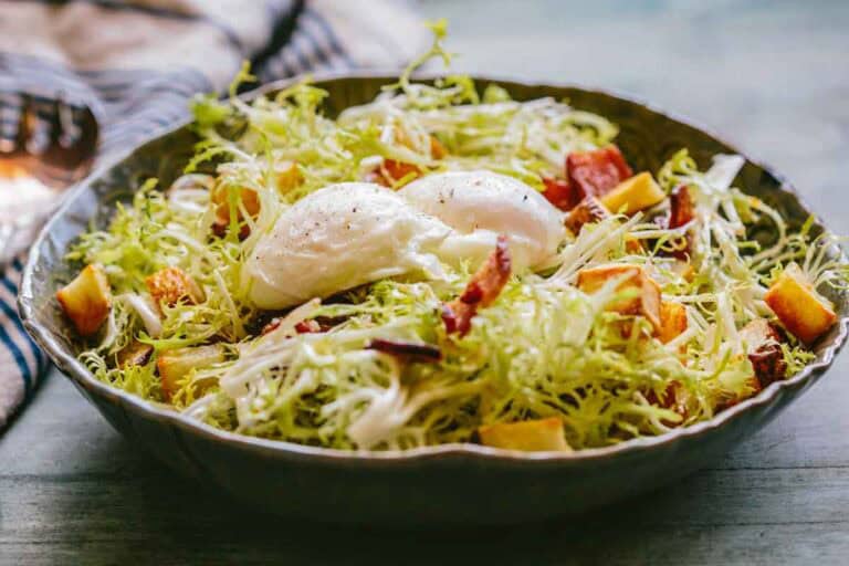 Side view of a Lyonnaise salad topped with two poached eggs nestled in a bed of frisée, with visible crispy bacon and potato croutons throughout.