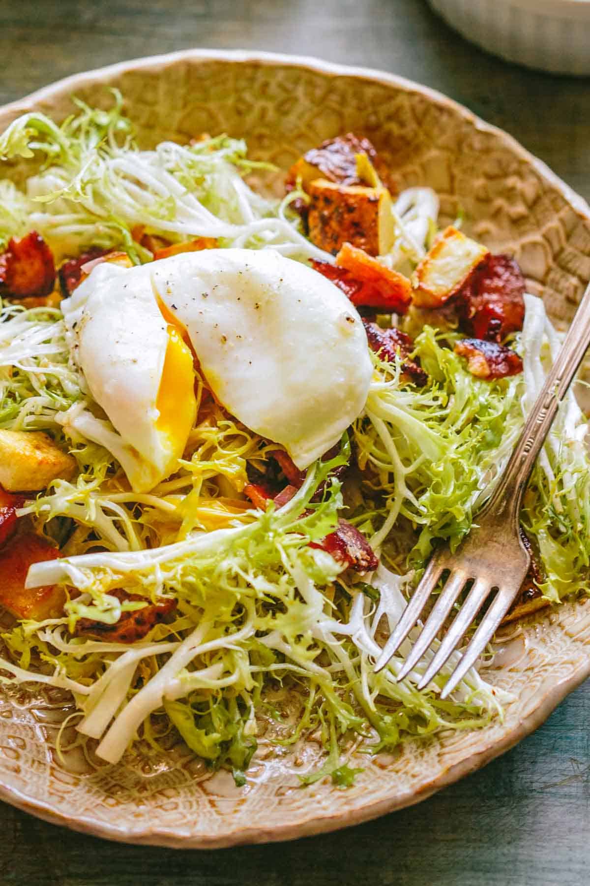 Close-up of a poached egg with its yolk beginning to ooze out over frisée lettuce, surrounded by crispy bacon and browned potato cubes.