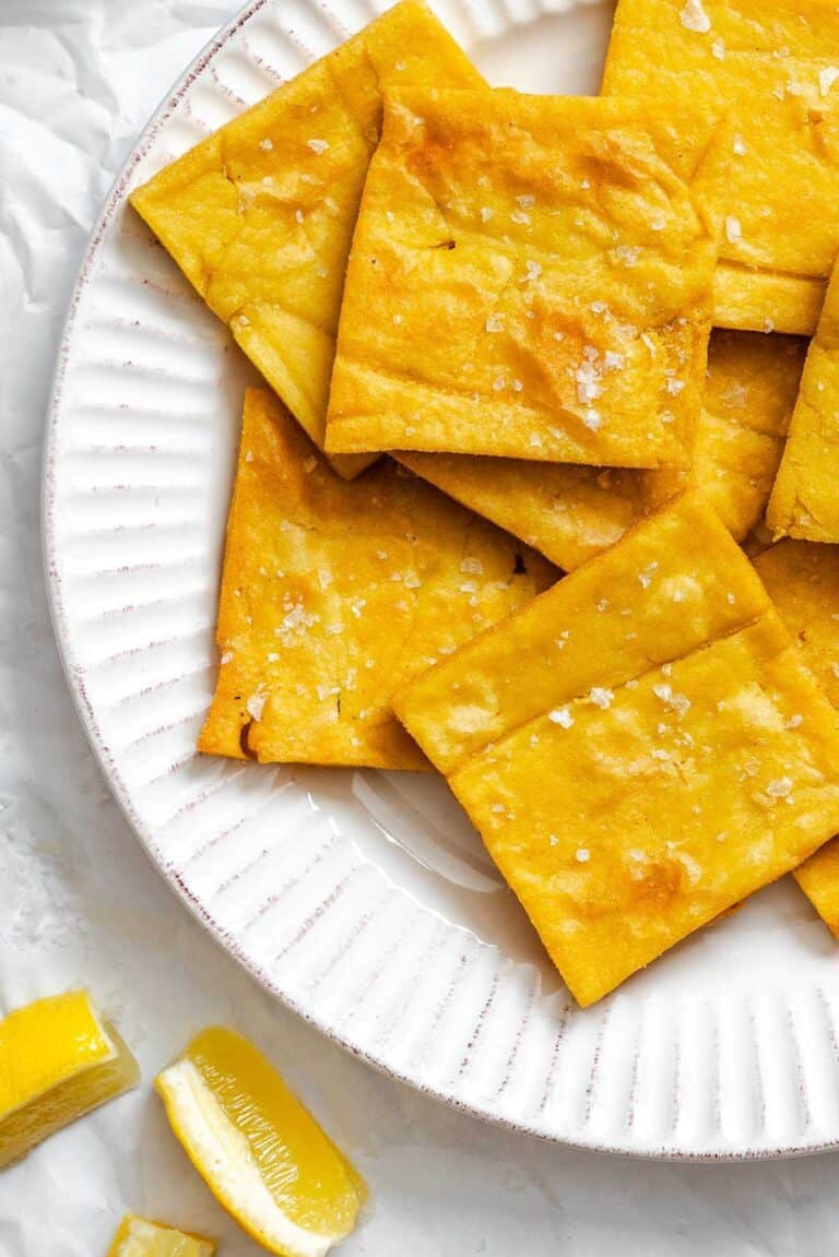 Golden, square-cut chickpea fritters (panelle) sprinkled with flaky sea salt, arranged on a white ridged plate with lemon wedges on the side.