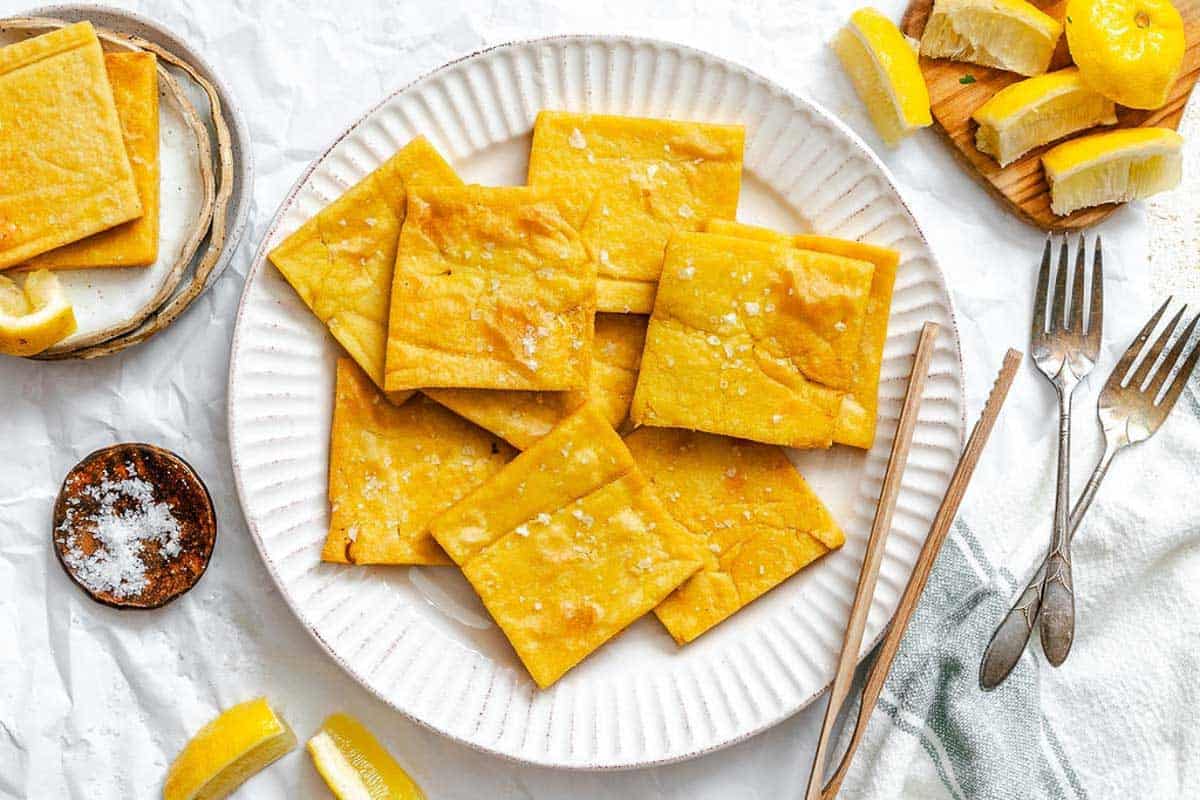 Top-down view of a serving platter filled with crispy panelle squares, surrounded by lemon wedges, salt, and utensils on a crinkled white paper backdrop.