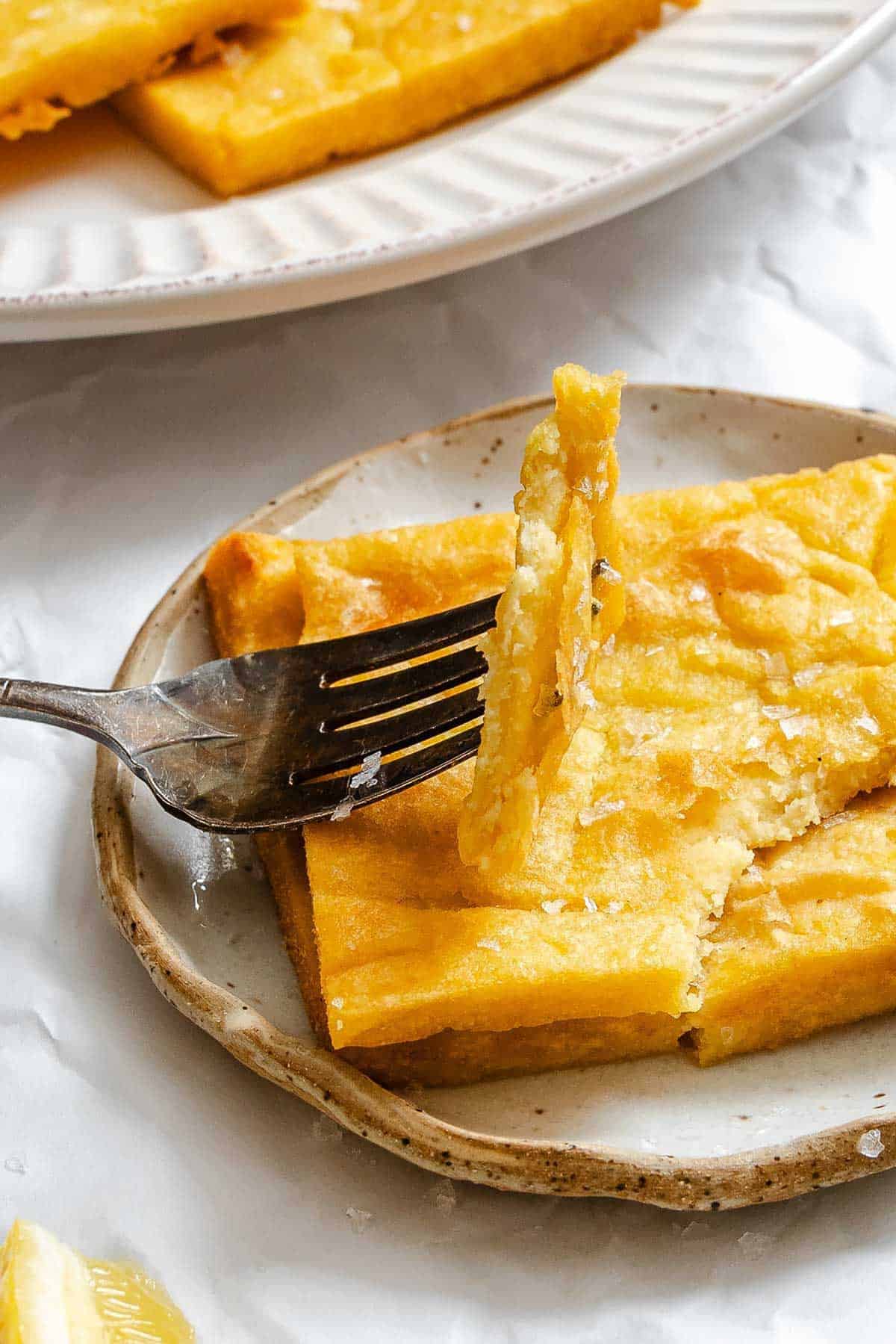A close-up shot of a fork lifting a tender piece of panelle to show its fluffy interior, with more fritters visible on a small ceramic plate below.