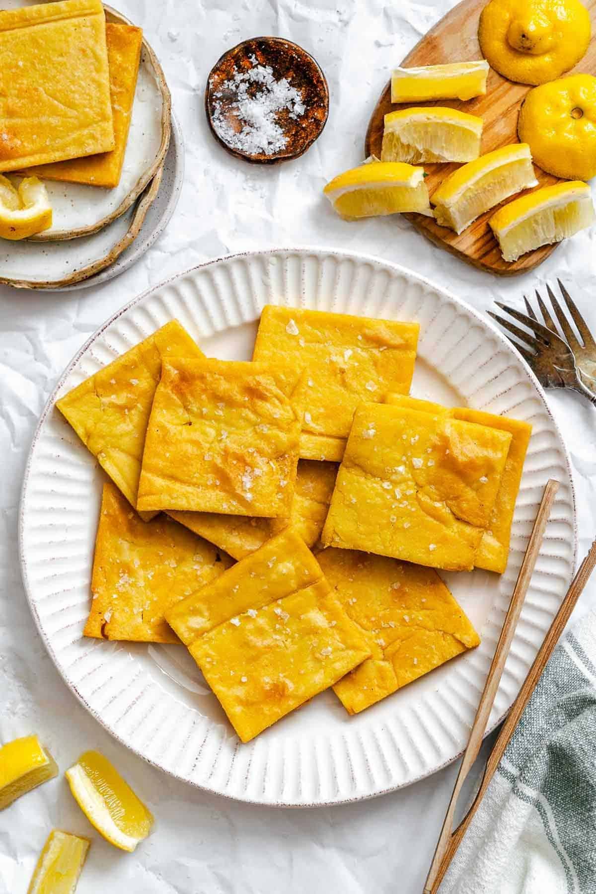 Bright overhead photo showing a plate of golden chickpea fritters surrounded by sliced lemons, salt, and cutlery—set for sharing or snacking.