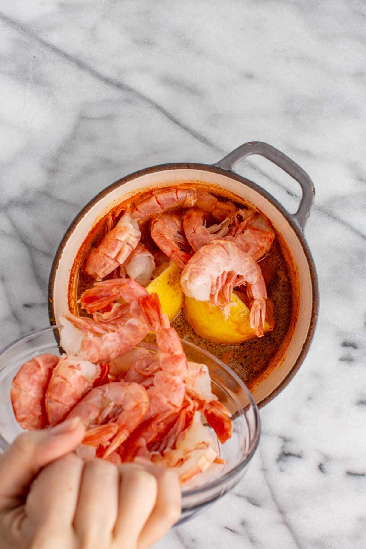 Raw shell-on shrimp being poured into the pot of spiced broth with halved lemons and seasonings floating on top.