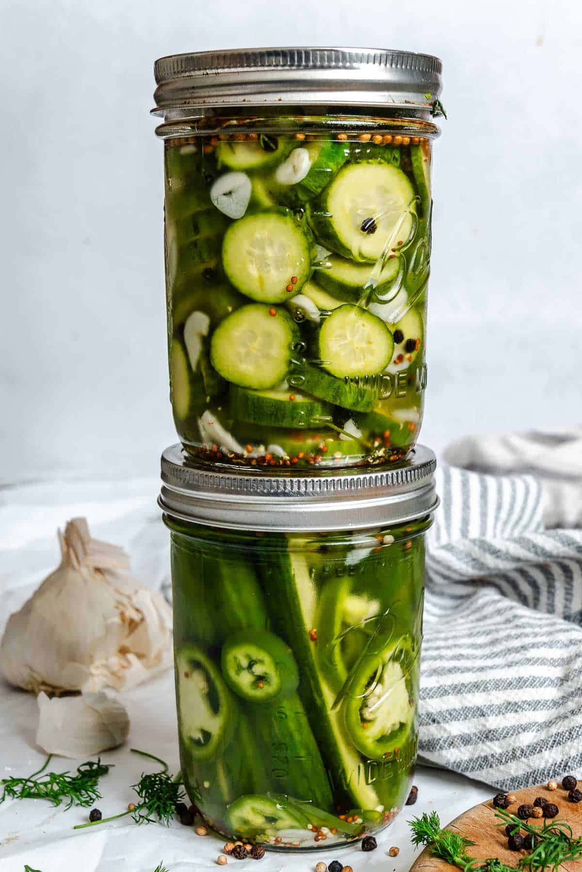 Two sealed jars of refrigerator pickles stacked on top of one another, filled with sliced cucumbers, jalapeños, garlic, and spices.