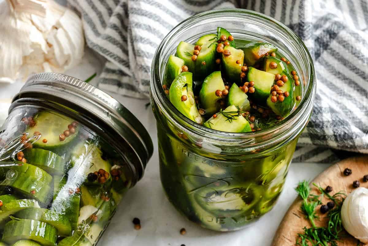 One jar lying on its side next to another upright, both filled with sliced cucumbers and mustard seeds in brine, with garlic and dill scattered around.