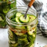 Fork inserted into an open jar of cucumber pickles, showing the slices submerged in brine with visible coriander and mustard seeds.