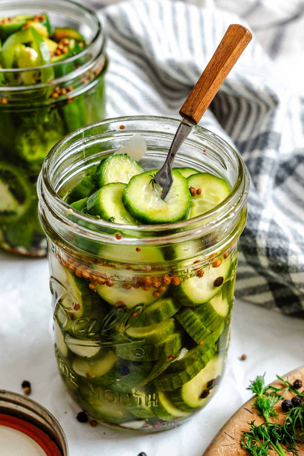 Fork inserted into an open jar of cucumber pickles, showing the slices submerged in brine with visible coriander and mustard seeds.