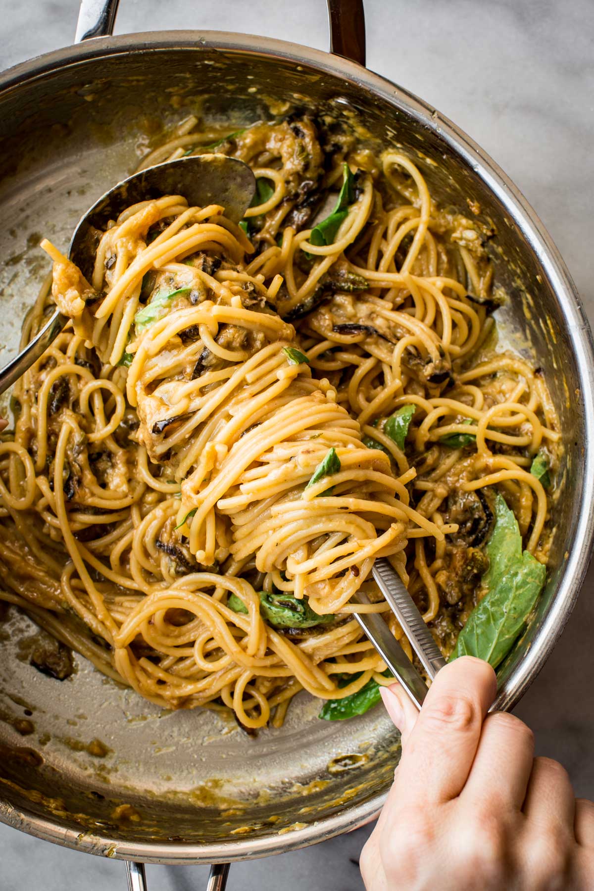 A close-up of spaghetti alla Nerano being twirled with tongs and a serving spoon in a stainless steel skillet. The creamy sauce clings to the pasta, with flecks of golden zucchini and fresh basil leaves visible throughout.