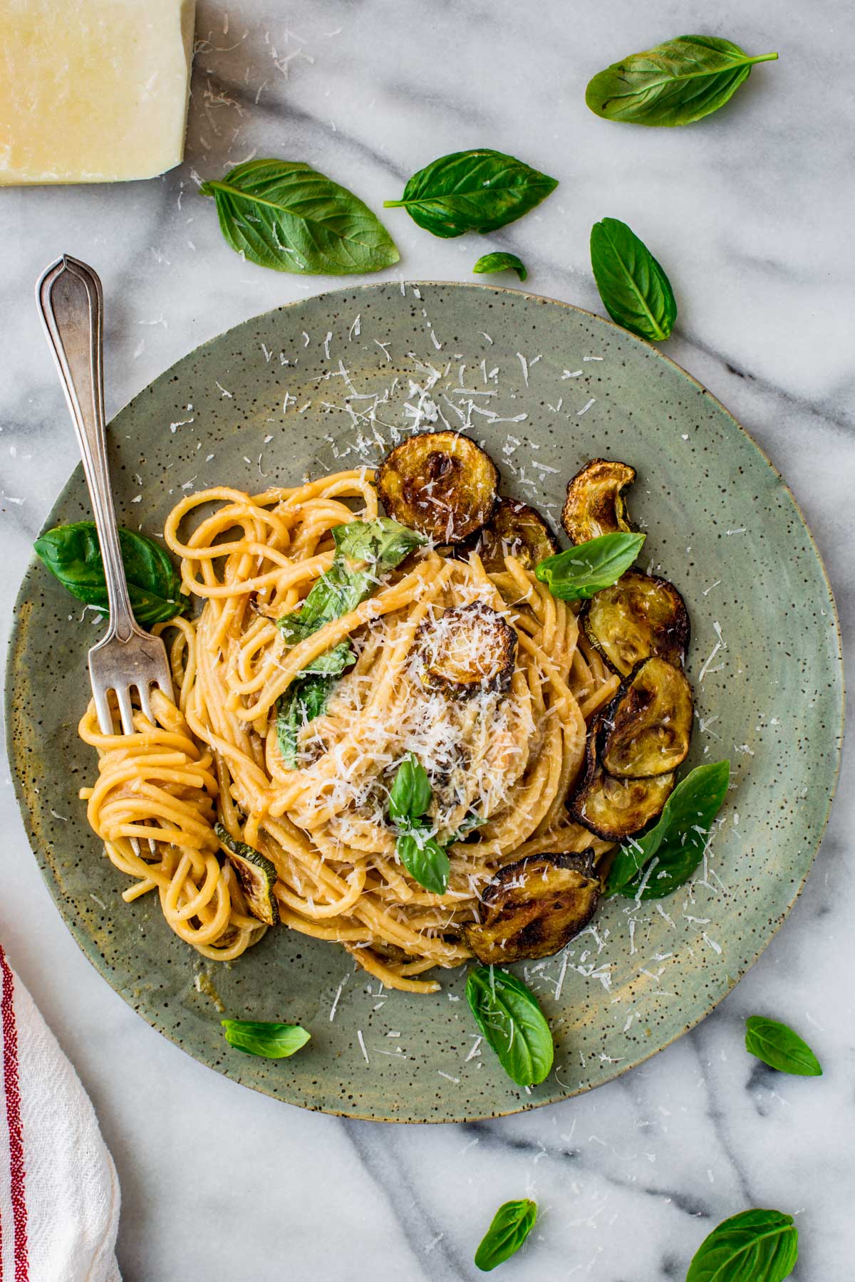 A rustic ceramic plate filled with a swirl of spaghetti alla Nerano topped with grated Pecorino Romano, crispy golden zucchini slices, and fresh basil leaves, set on a marble background with a fork tucked into the side.