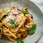A detailed view of spaghetti alla Nerano on a speckled ceramic plate, showing the rich, creamy sauce, shreds of Pecorino Romano cheese, blistered zucchini chips, and vibrant green basil leaves.