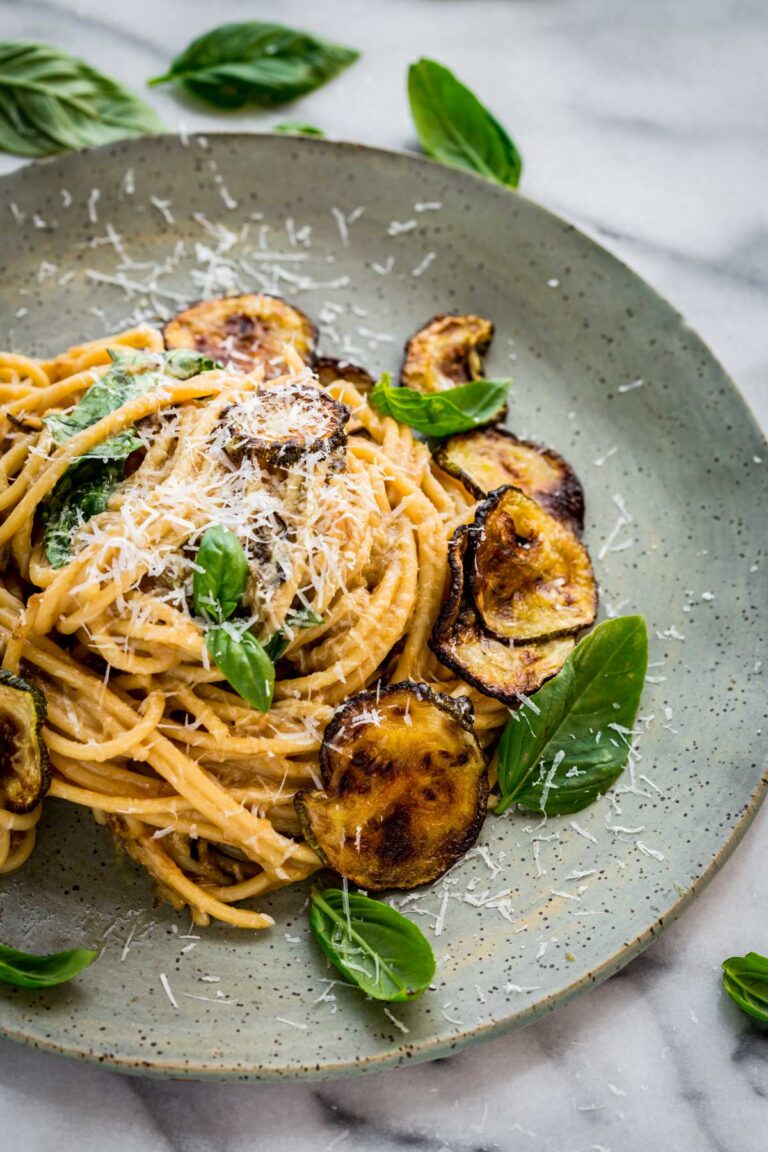 A detailed view of spaghetti alla Nerano on a speckled ceramic plate, showing the rich, creamy sauce, shreds of Pecorino Romano cheese, blistered zucchini chips, and vibrant green basil leaves.