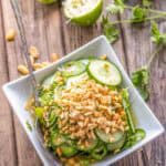 Overhead view of Thai cucumber salad in a square white bowl on a rustic wooden surface. The salad is topped with crushed peanuts and garnished with cilantro. Around the bowl are scattered peanuts, cucumber halves, lime halves, and cilantro sprigs for a casual, fresh presentation.