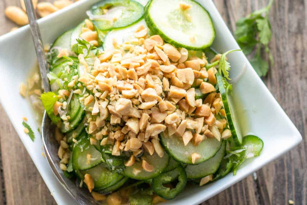 A close-up of a Thai cucumber salad served in a white square bowl. Thin slices of cucumber are layered with fresh cilantro, thinly sliced green chiles, and a generous mound of chopped peanuts on top. A spoon rests inside the bowl.