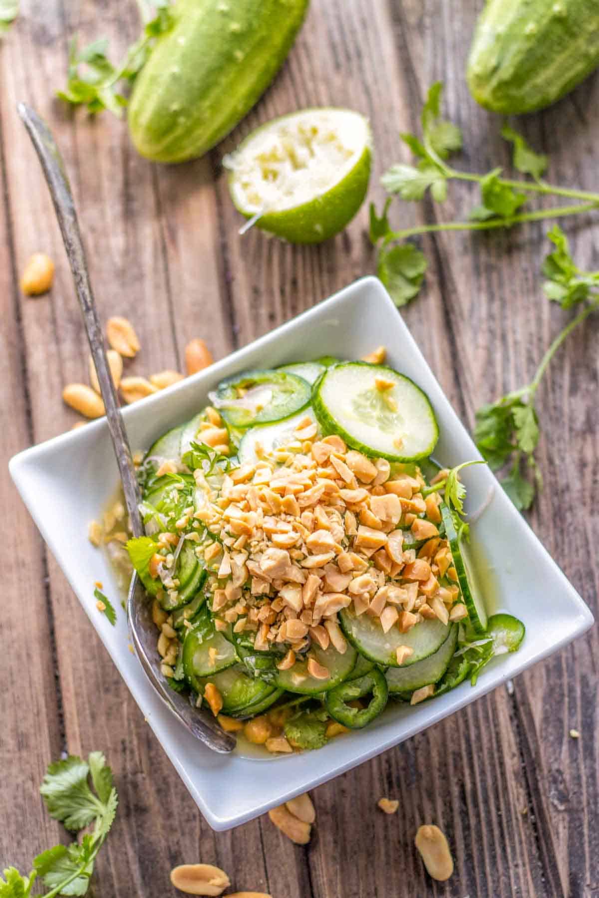 Overhead view of Thai cucumber salad in a square white bowl on a rustic wooden surface. The salad is topped with crushed peanuts and garnished with cilantro. Around the bowl are scattered peanuts, cucumber halves, lime halves, and cilantro sprigs for a casual, fresh presentation.