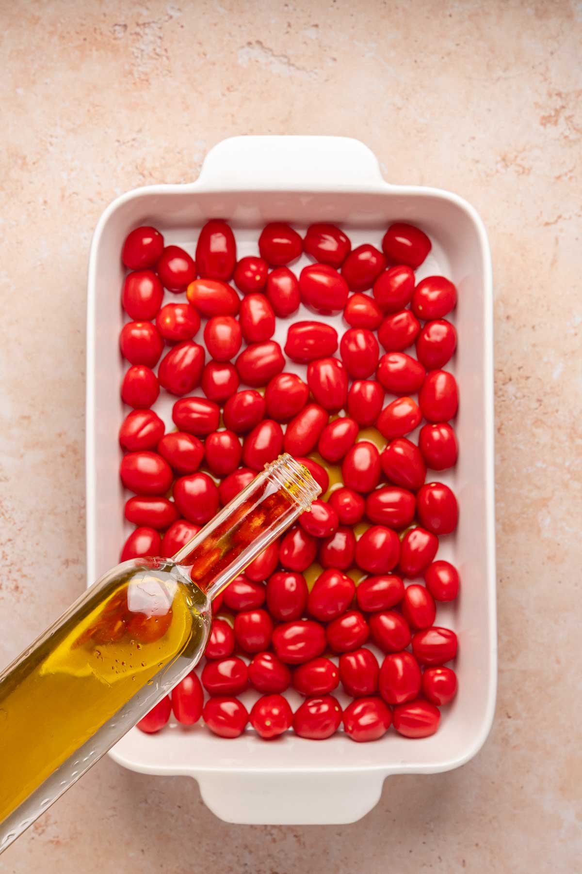 Bottle of olive oil being poured over cherry tomatoes in a white baking dish.