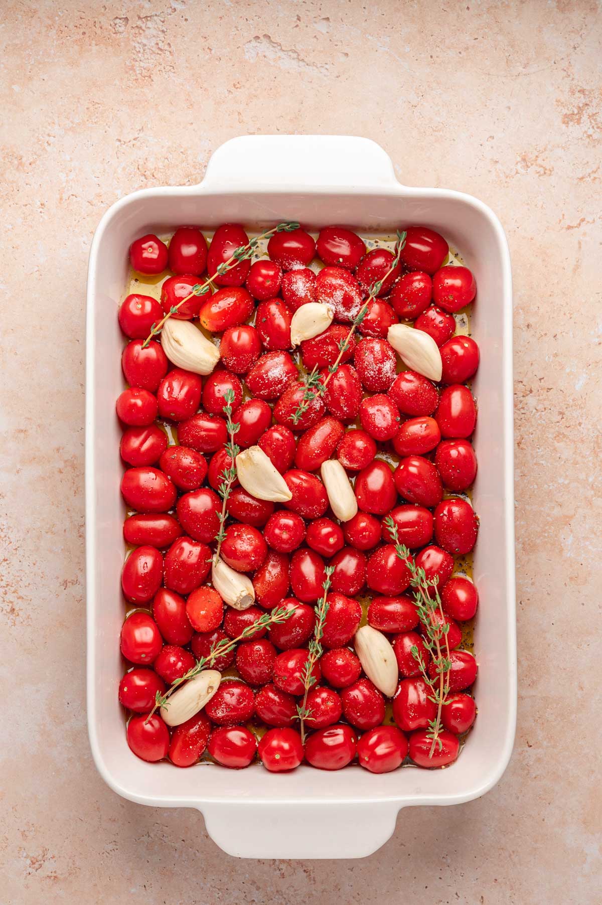 Cherry tomatoes topped with garlic cloves and thyme sprigs, ready for roasting.